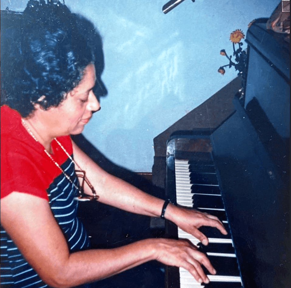 Mara Passos tocando piano com expressão acolhedora e luzes aconchegantes